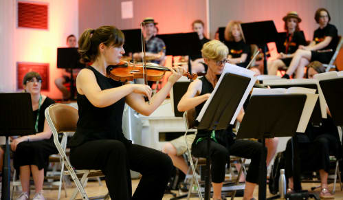 A female violinist sits at the front of an orchestra of adult professional musicians and young people
