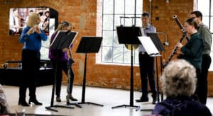 Five musicians - all playing wind instruments - stand in a circle around music stands. They are indoors, stood in front of a brick wall with a large window.