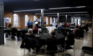 A large room with brick walls and tall windows. A band of five musicians perform in front of an audience sat around tables.