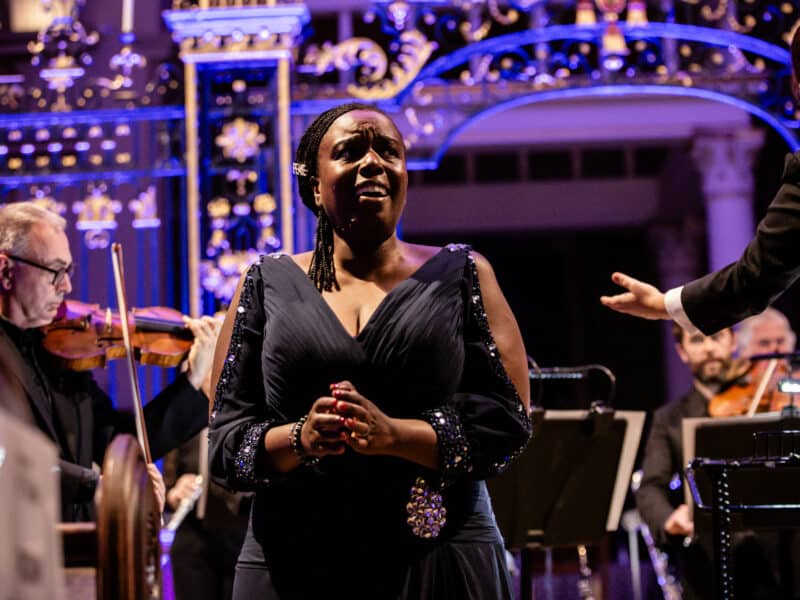 Nadine Benjamin wearing a sparkly dress smiling and looking over to the side while people play instruments in the background