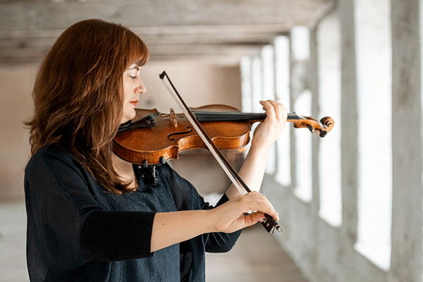 A woman in black concert dress is in a room white walls playing violin. She faces right towards the windows.