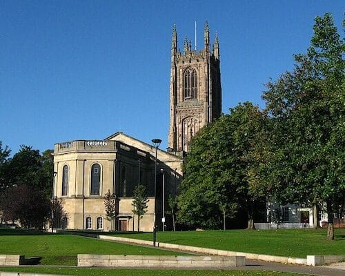 Picture shows Derby Cathedral set against a blue sky, with large trees in the foreground.