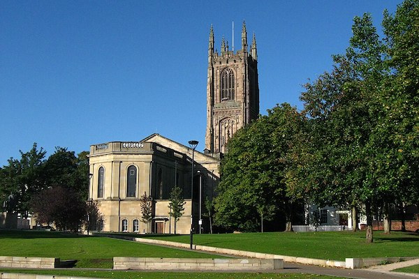 Picture shows Derby Cathedral set against a blue sky, with large trees in the foreground.