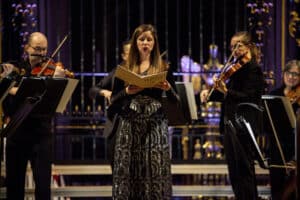 A woman holds a musical score and sings, flanked by two violinists.