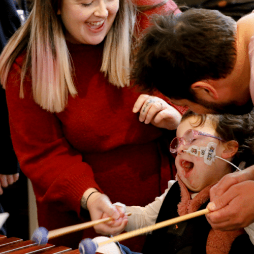 A woman and a man helping a child with additional needs and a feeding pipe enjoy playing the glockenspiel