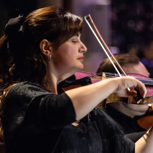 Sophie Rosa plays violin. She has dark hair half tied up with a bow and a fringe. She is wearing evening concert blacks. There is another musician in the background.