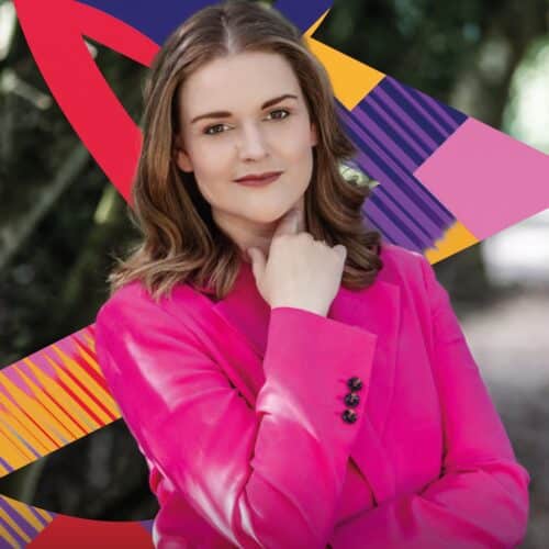 Image shows soprano Jessica Cale in a pink jacket, standing in the woods